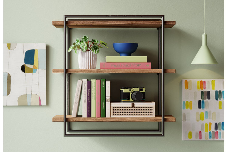 Three-tiered wood and metal wall shelf mounted on a green wall beside two abstract prints and used to display books and decor.