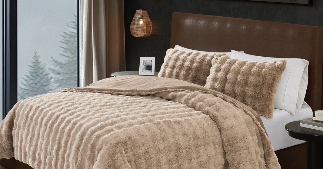 A cozy bedroom featuring a tan, textured faux fur comforter set with matching shams on a bed with a dark leather headboard, set against a window with a wintry forest view.