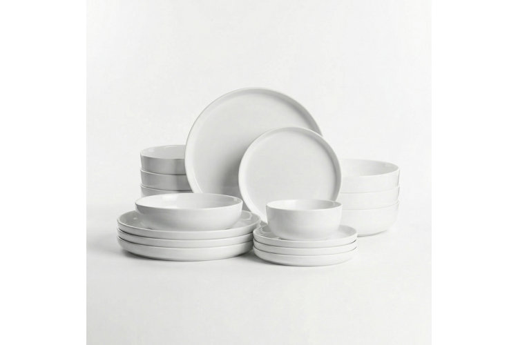 A product shot of a minimalist all-white ceramic dinnerware set isolated on a white background. The collection is organized into neat stacks and includes large porcelain dinner plates, smaller salad plates, wide shallow bowls, and deep cereal bowls, all featuring clean lines and a smooth matte finish.