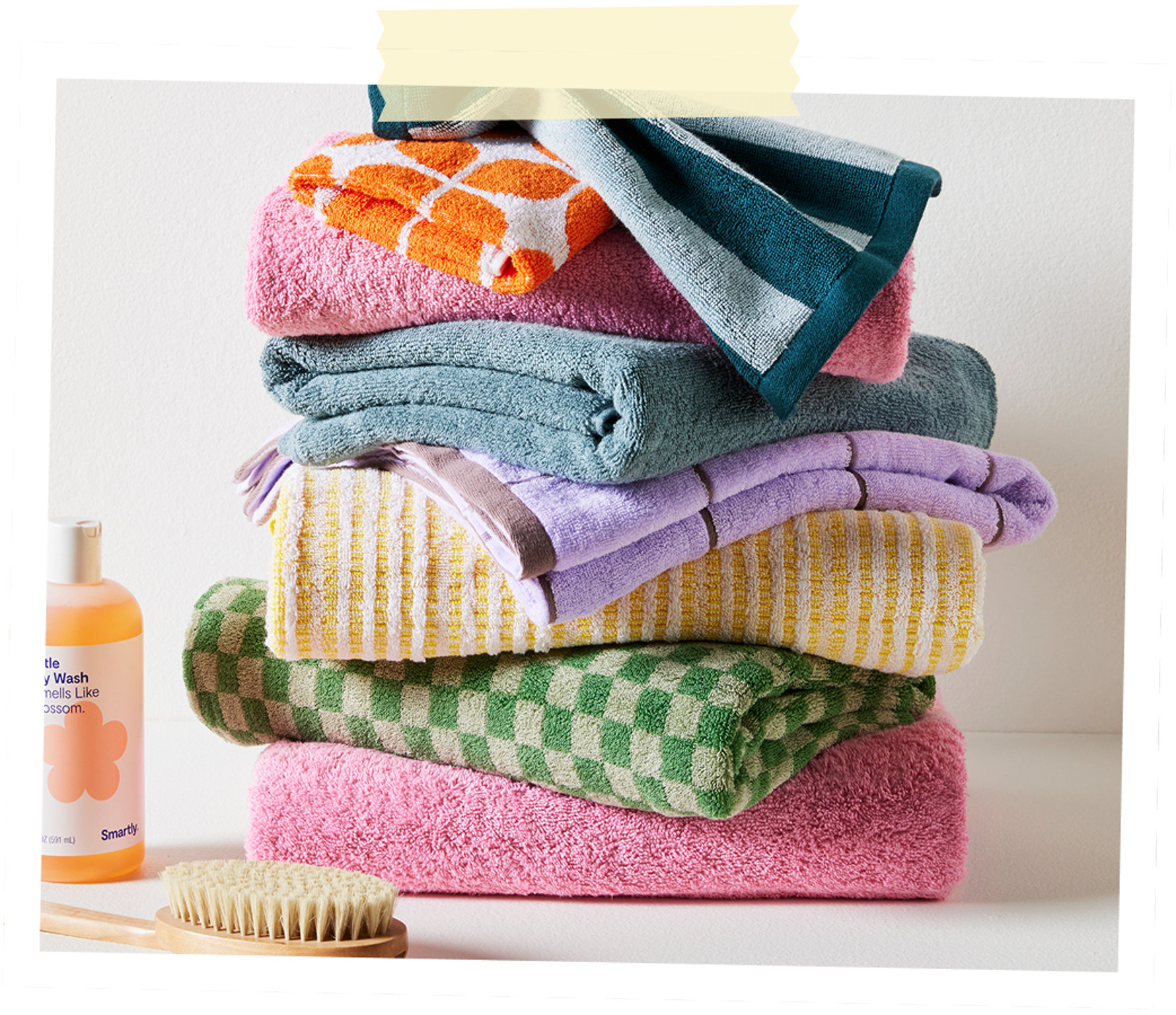 A tall stack of colorful, patterned bath towels next to a bottle of body wash and a wooden bath brush.