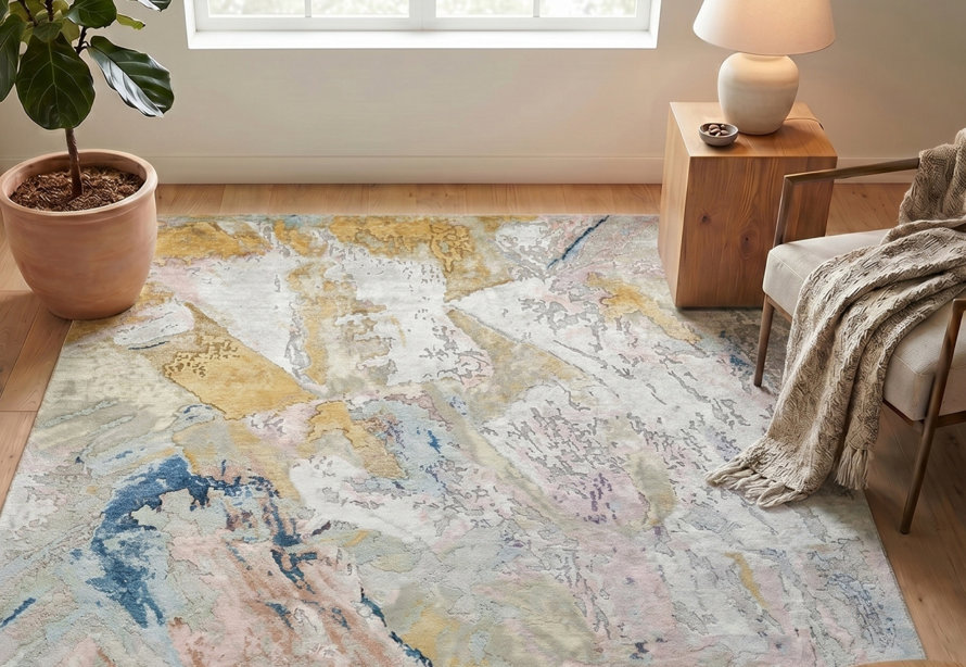 An abstract area rug with mottled patches of gold, blue, light pink, and ivory displayed in a bright, modern living room with a houseplant, wood side table, and armchair.