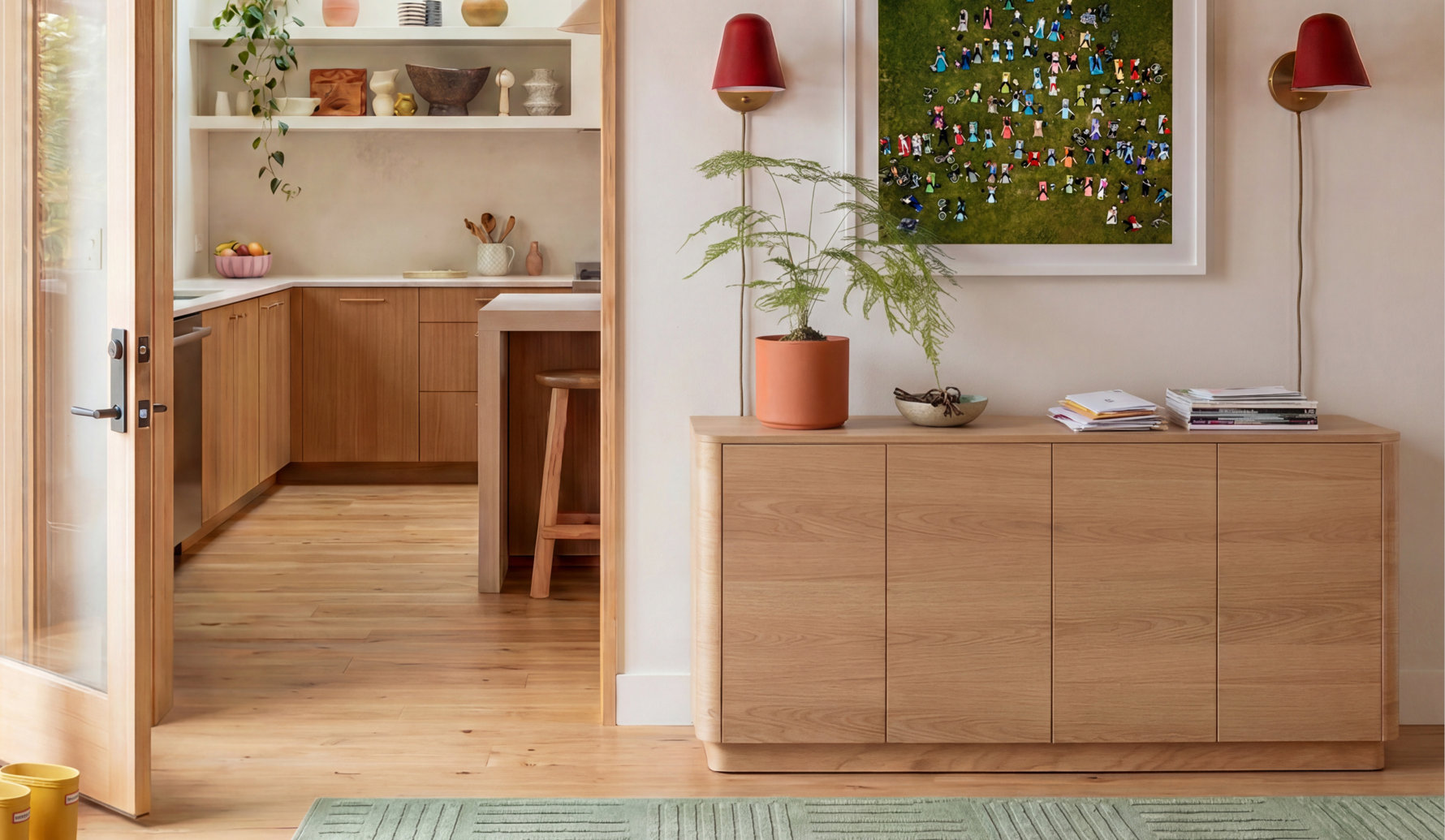 A modern wooden sideboard with rounded edges sits in a brightly lit entryway, topped with a potted plant, a bowl, and a stack of mail, framed by two red wall sconces and an art print, with a view into a warm kitchen featuring light wood cabinetry.
