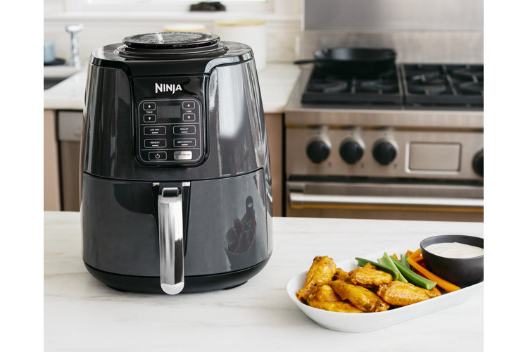 Black air fryer with a white condiment serving dish filled with chicken wings and other finger foods