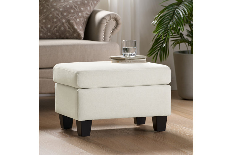 A small square storage ottoman upholstered in an off-white textured fabric, featuring a thick cushioned top and four short, dark-brown tapered block feet, styled in a living room next to a potted plant and a beige sofa
