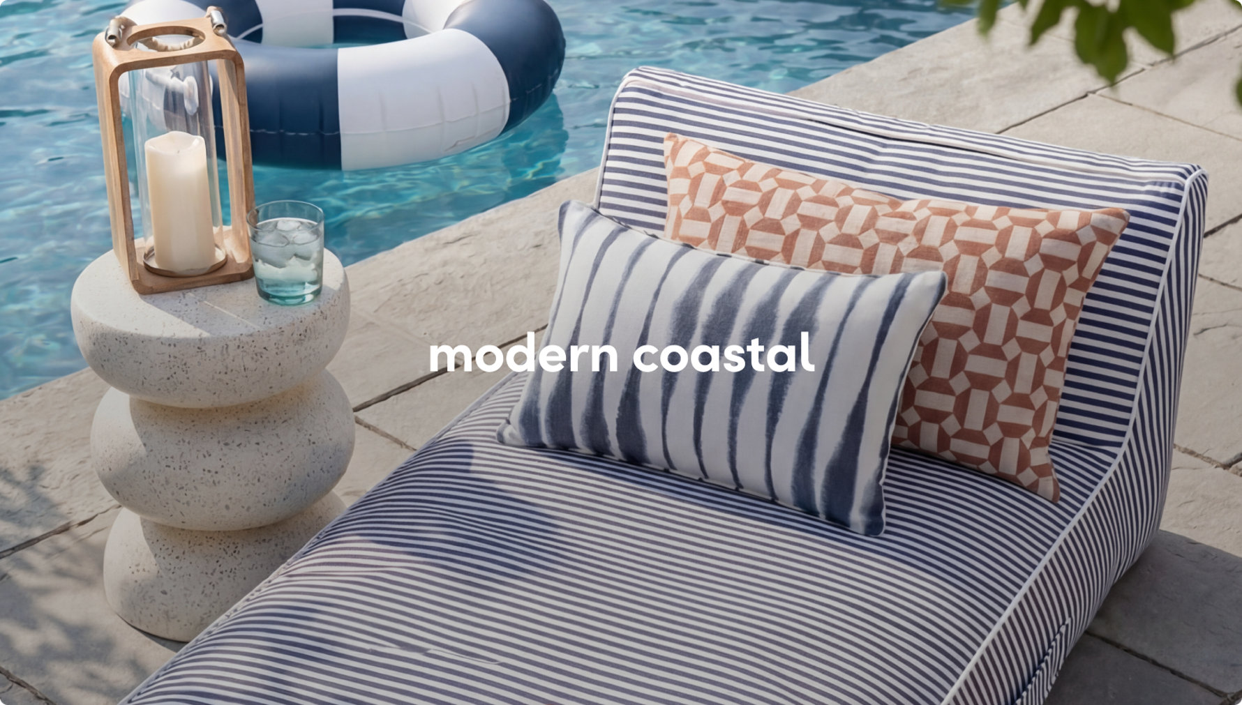 A striped outdoor chaise lounge with blue and tan pillows next to a stone side table and a candle lantern by a pool.