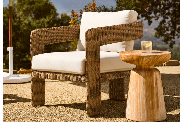 Woven outdoor lounge chair with white cushions beside a round wooden side table on a sunlit patio