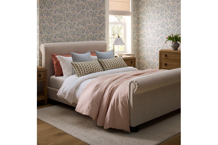 where to place a bed in a bedroom cream colored upholstered sleigh bed with orange striped bed set and blue and orange throw pillows blue patterned wallpaper and two light wood night stands with tabletop lamps and a window on either side of the bed