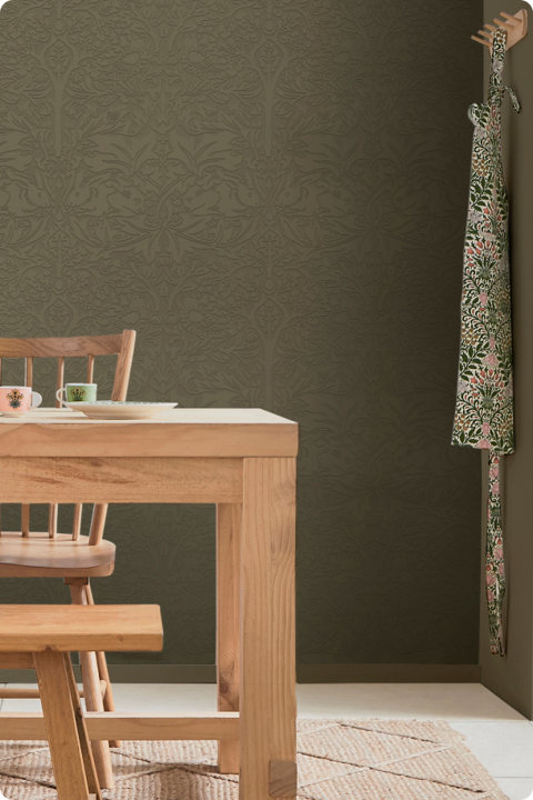 Dining room with olive green embossed wallpaper featuring a subtle rabbit and floral pattern, a light wood dining table and chairs, and a floral apron hanging on the wall.
