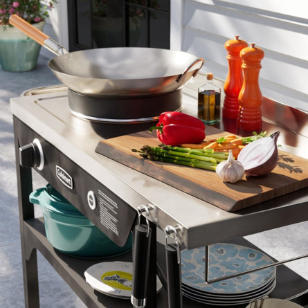 Vegetables on a cutting board next to a wok on an outdoor kitchen island.