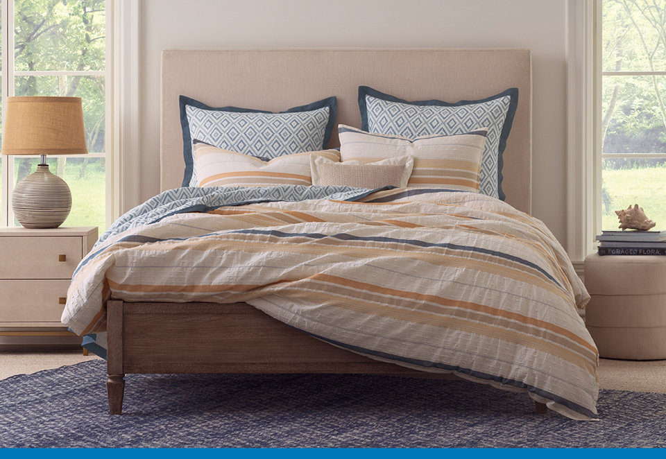 A queen bed with a tufted beige headboard, dressed in a white, blue, and orange striped duvet and geometric patterned pillows, set in a bright bedroom with two nightstands and windows on either side.