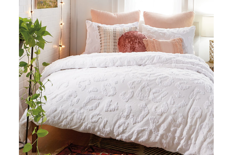 A bed with white textured bedding that has a subtle cheetah print paired with pink throw pillows and beside a pothos plant.